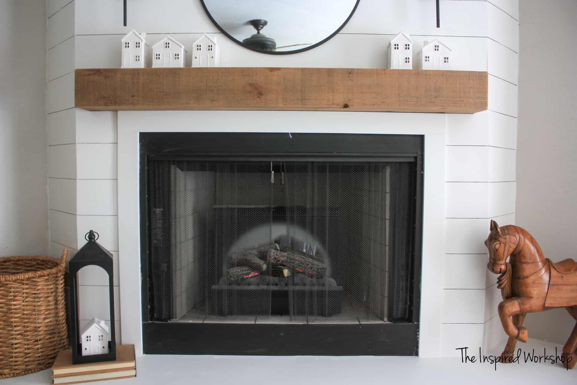 white shiplap Fireplace up close with a cedar mantle, a wooden horse, books, a lantern and white houses to decorate the fireplace