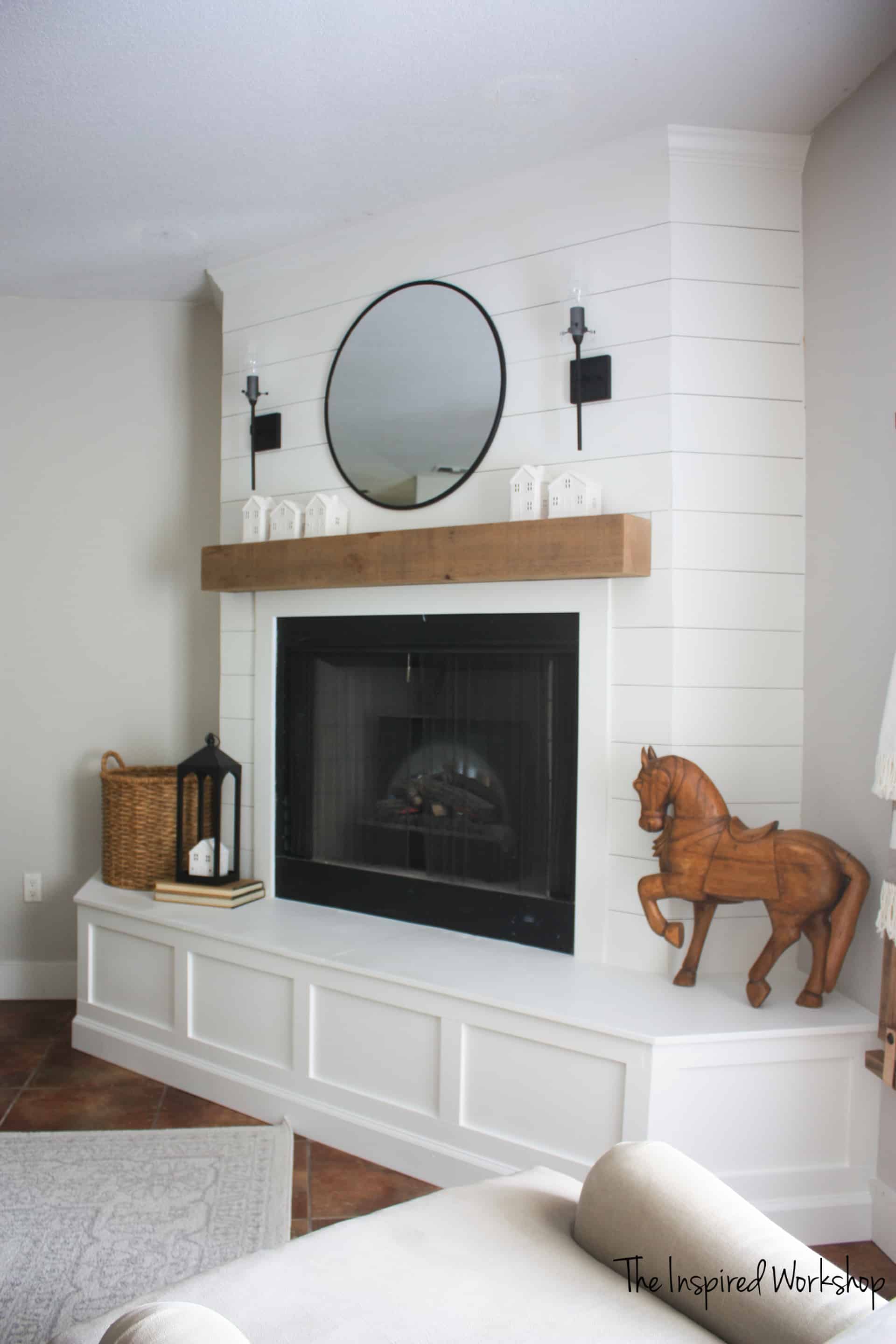 Finished picute of the Fireplace Makeover shown from the side all white fireplace with round mirror above the cedar mantle and wall mouted lights. a wooden horse and lantern with wicker basket decorate the hearth