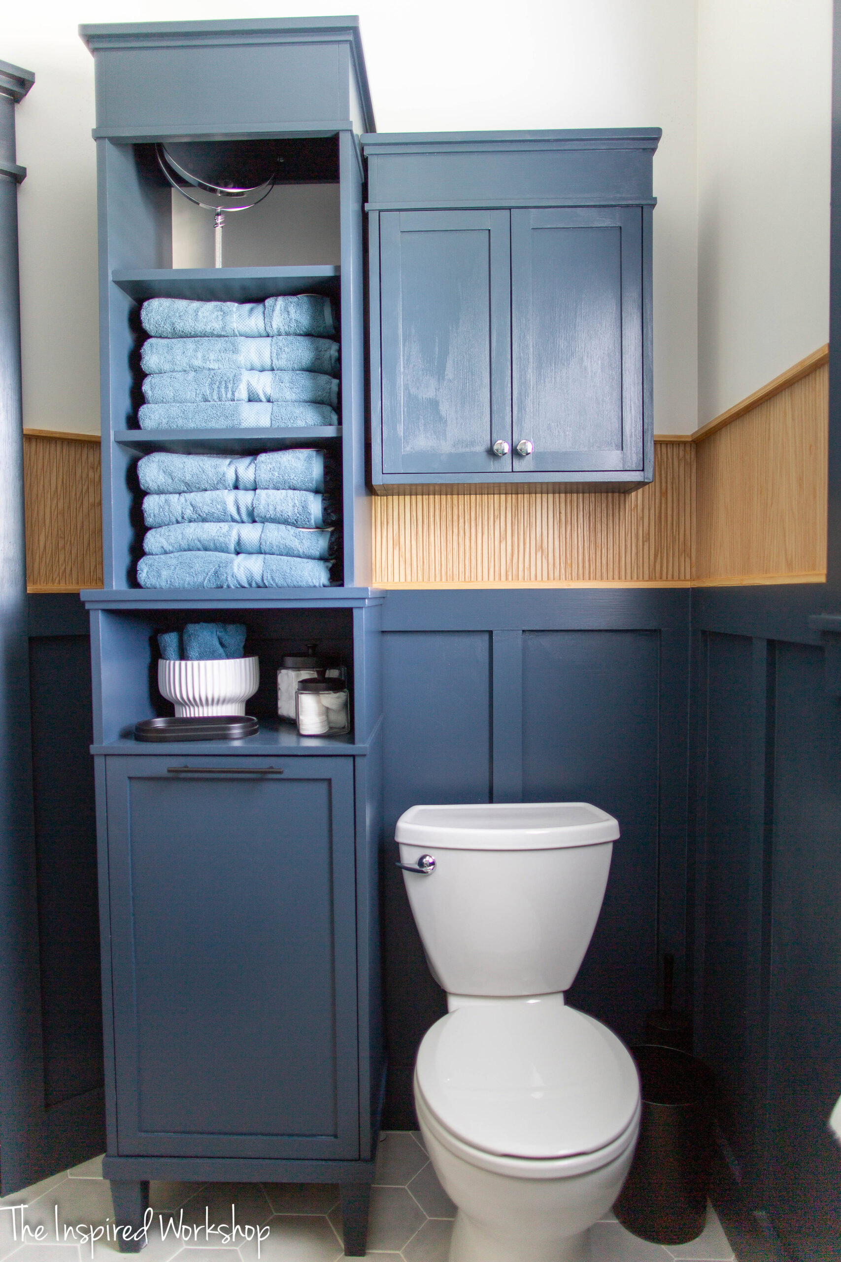 Navy blue linen tower with open shelves on top and a toilet to the right with a navy blue cabinet above the toilet.